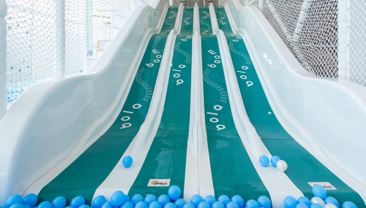 A brightly colored indoor playground slide with multiple lanes and safety nets, surrounded by scattered blue and white plastic balls at the bottom. Bolaboom adds an extra layer of fun!