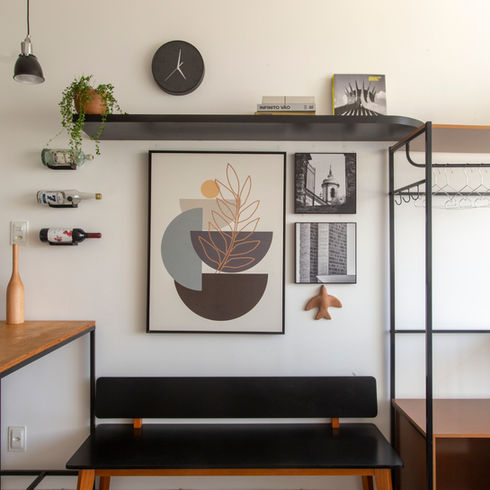 Foto - Laranjeiras 336. Banco em madeira com tampo em encosto na cor preta, quadros de pinturas e imagens acima do banco, e acima dos quadros uma prateleira preta com vaso de planta, um relógio analógico preto. Ao lado direito cortada da imagem, um armário aberto. Ao lado esquerdo cortada da imagem uma Bancada de serralheria de estrutura em perfil de aço 2x2cm na cor preta com tempo em madeira. Teto em rebaixo de gesso na cor branca, com pendentes sobre a bancada e spots sobre o armário, parede na cor branca e piso cerâmico cinza claro.