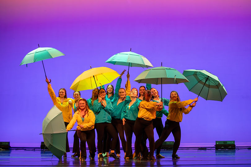 Group of dancers in tap shoes pose center stage. They are wearing a mixture of teal and yellow raincoats, and holding five teal umbrellas. One yellow umbrella is being held by the lead dancer. The background is a blend of blue and purple lighting.