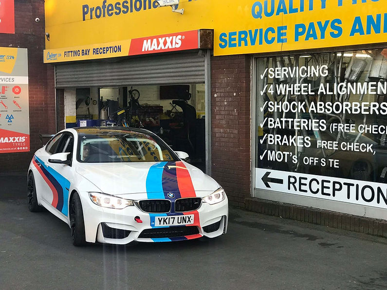 A white BMW with blue and red racing stripes is parked outside a garage with signs for services like wheel alignment and brakes