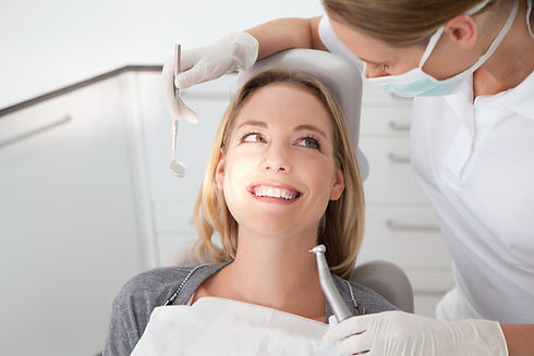 A woman smiles while sitting in a dental chair, looking at a dentist holding tools