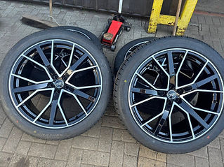 Two glossy, black alloy wheels with silver accents are stacked on a tiled floor