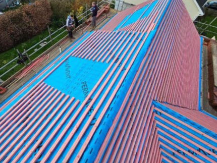 A rooftop in progress covered with blue and red striped waterproofing materials, with workers standing on scaffolding