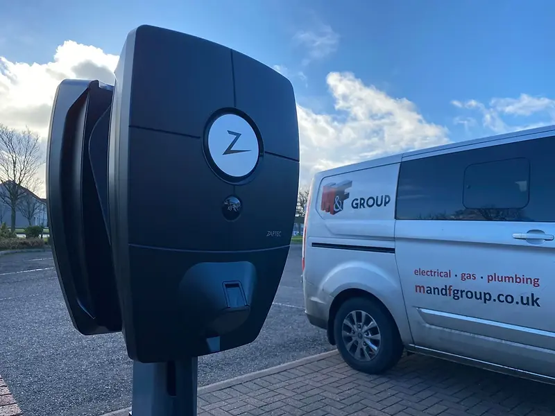Close-up of an electric vehicle charging station with a circular logo, positioned beside a white van with branding for an electrical and plumbing company