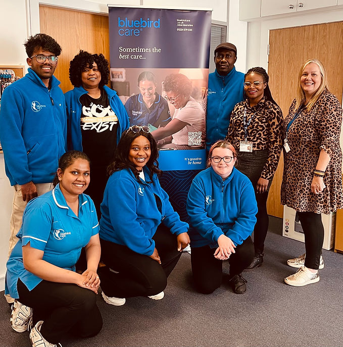 A group of eight people, mostly wearing blue uniforms, stand smiling in an office