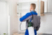 A pest control worker in a blue and gray uniform sprays inside a kitchen cabinet, holding a canister
