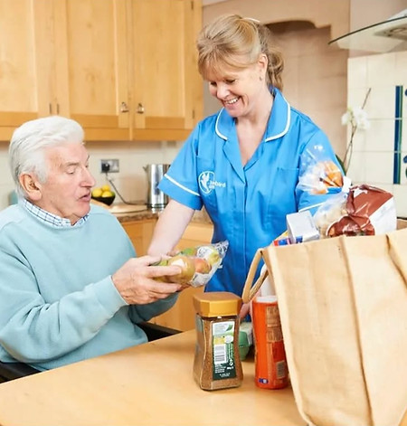 A caregiver in a blue uniform helps an elderly man in a kitchen, sharing groceries from a bag