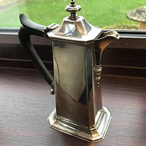 A polished silver coffee pot with an ornate spout and black handle sits on a wooden windowsill, reflecting light