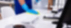 A person wearing blue gloves cleans an office desk with a yellow cloth and spray bottle