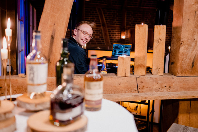 Hochzeits-DJ Alexander Stahl aka STEEL auf einer Hochzeit im Spargelhof Schulte-Scherlebeck in Herten. Er wird schräg von hinten fotografiert und lächelt in die Kamera. Im Vordergrund sind verschiedene Whiskeyflaschen zu erkennen. Foto: Maximilian Ishorst.