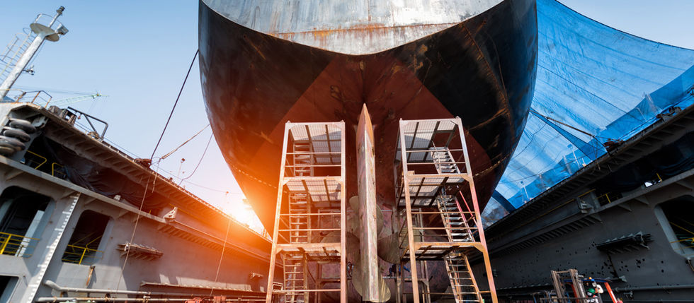 Close-up view of a ship in dry dock undergoing dismantling or maintenance, with scaffolding and workers visible beneath the hull, representing end-of-life vessel preparation.
