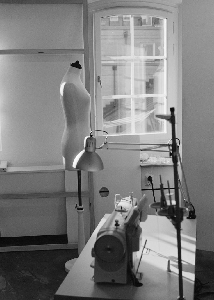 A black-and-white photograph of a minimalistic sewing studio. The image features a dress form standing near a large arched window with soft daylight streaming through. In the foreground, there's a heavy-duty sewing machine on a worktable, accompanied by adjustable task lamps. The scene captures a quiet, artistic atmosphere, evoking the timeless elegance of fashion design in progress.