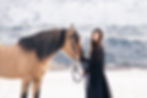 a girl wearing a long black dress standing in snow with a mountain in the background, petting her buckskin mustang horse