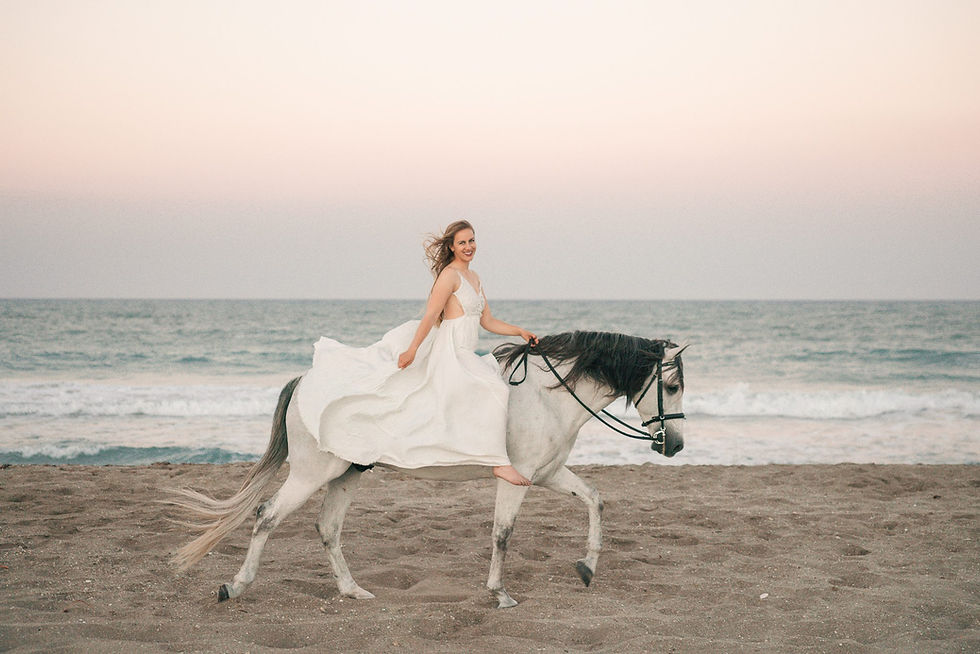 a blonde girl wearing a white dress sitting on her grey andalusian horse as they walk along the beach in Florida