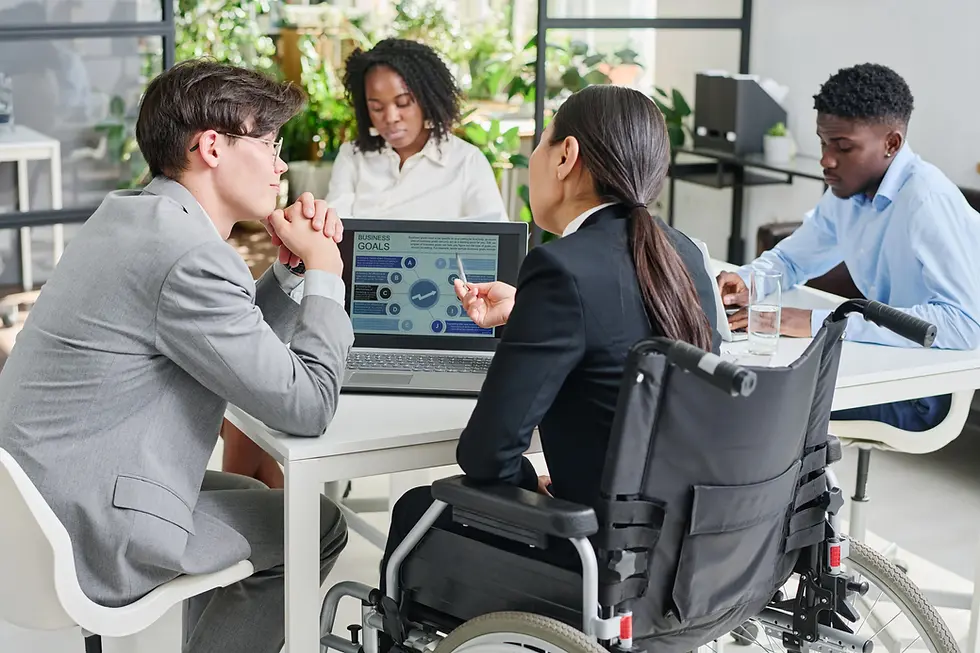 Four people are gathered around a table in an office setting. A woman in a black suit sits in a wheelchair, pointing at a laptop screen displaying business goals. Two men, one in a light grey suit and another in a blue shirt, are listening attentively. Behind them, a woman with curly dark hair sits at the table, also engaged in the discussion. A glass of water is on the table. The scene is bathed in natural light. This image can be used for projects that focus on business, collaboration, inclusivity, and workplace dynamics.