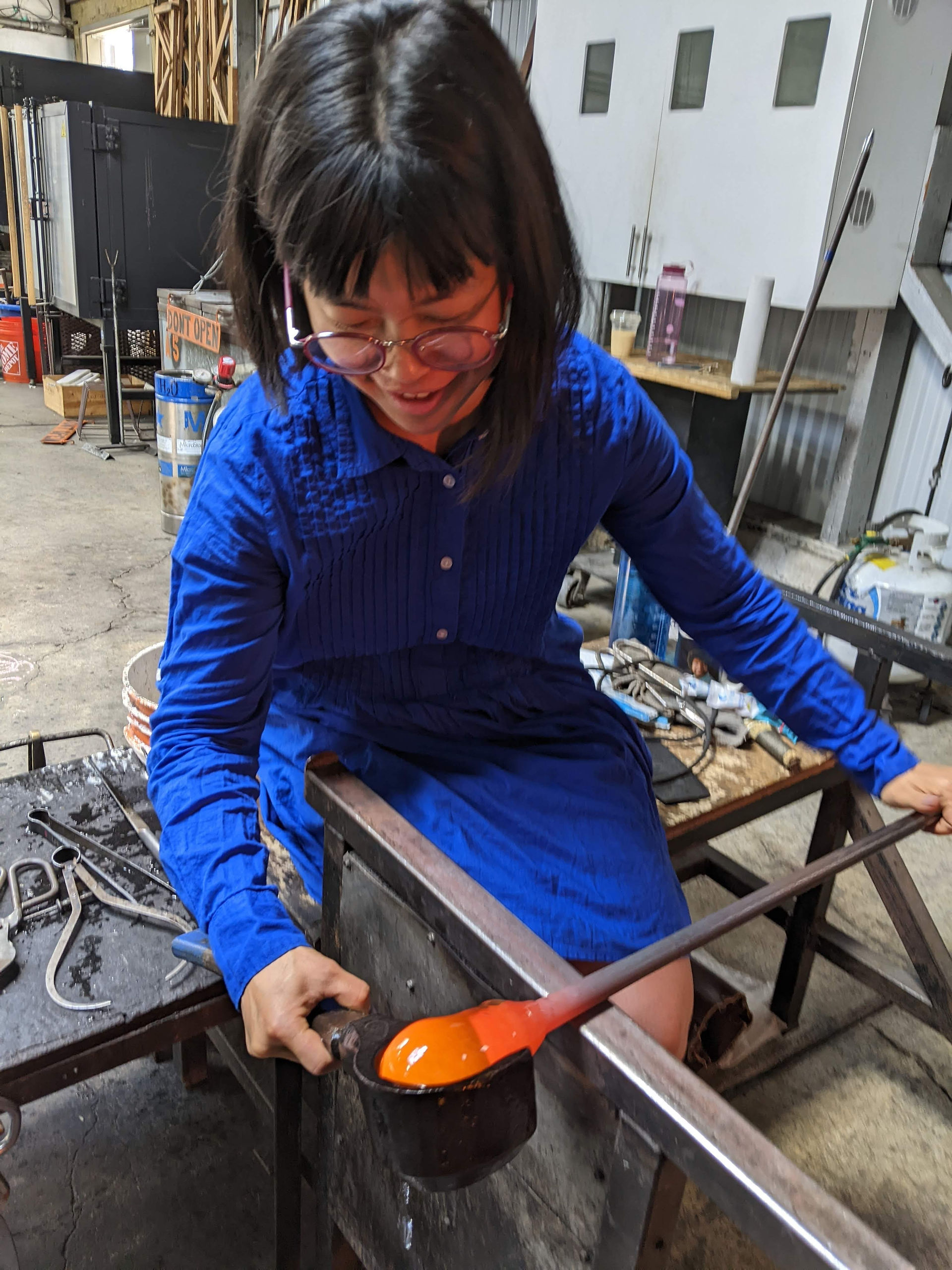 Maria in a bright, royal blue cotton dress and blowing glass at the bench. She is looking down at the glass bubble and shaping it with a block.