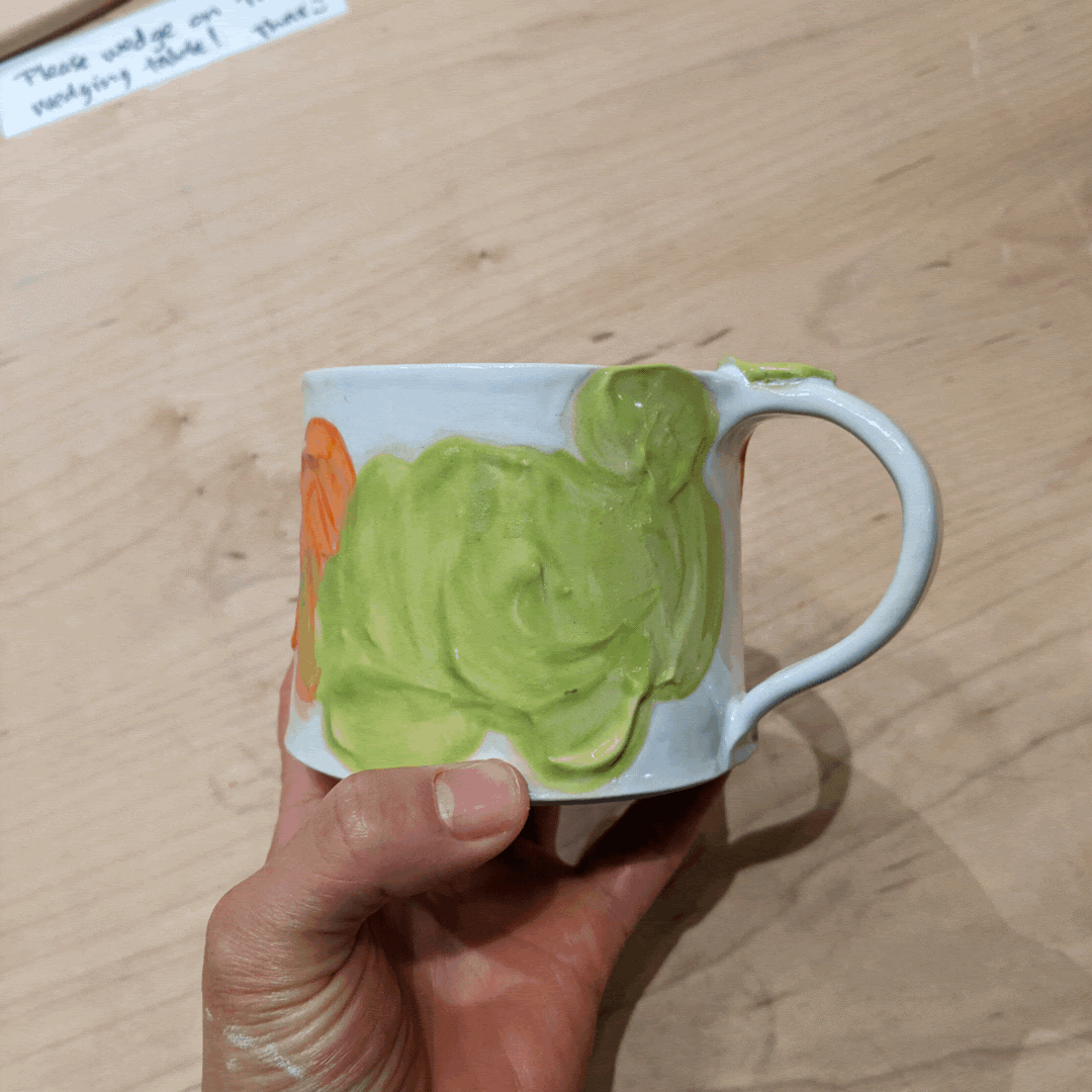 White ceramic mug with dimensional slip decorations, overlaid with bright green and orange paint. The glaze is reminiscent of abstract flora.