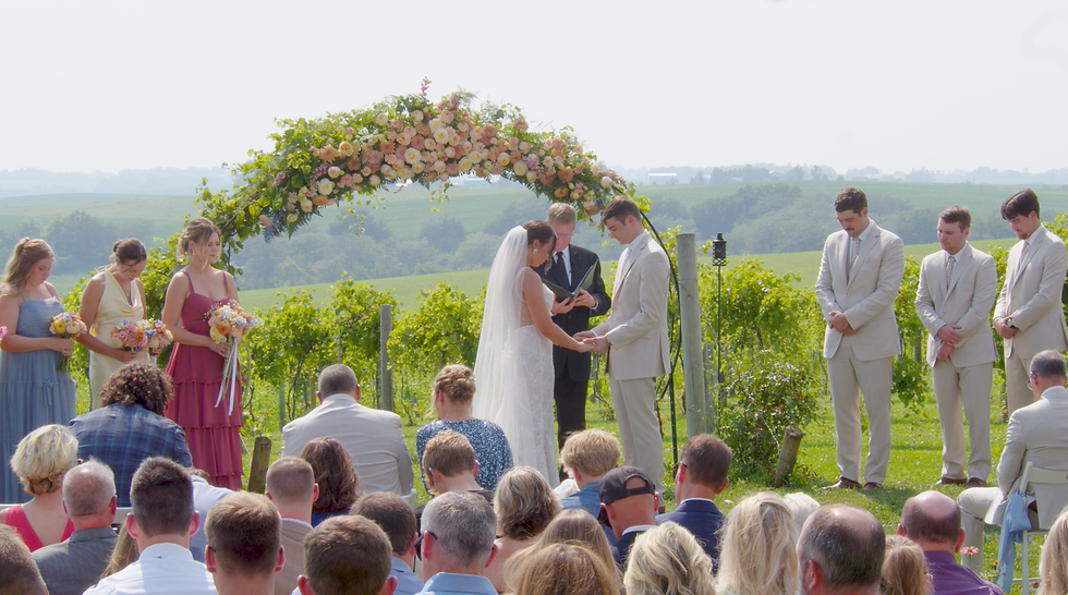 Outdoor wedding ceremony under a flower arch, bride and groom exchanging vows as bridesmaids and groomsmen stand nearby captured by an Eastern Iowa wedding videographer.