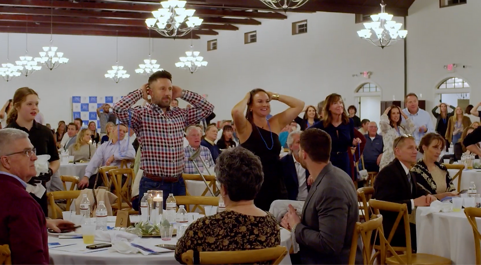 Guests at a banquet hall stand with hands on their heads during a lively game under bright chandeliers captured by an Eastern Iowa wedding videographer.