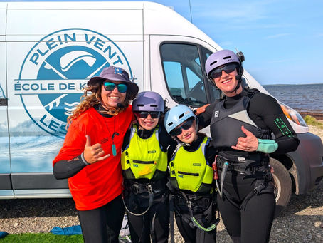 Une instructrice de Plein Vent les Îles sourit avec une mère et ses deux enfants équipés de combinaisons, gilets de sauvetage et casques de communication devant le van de l'école.