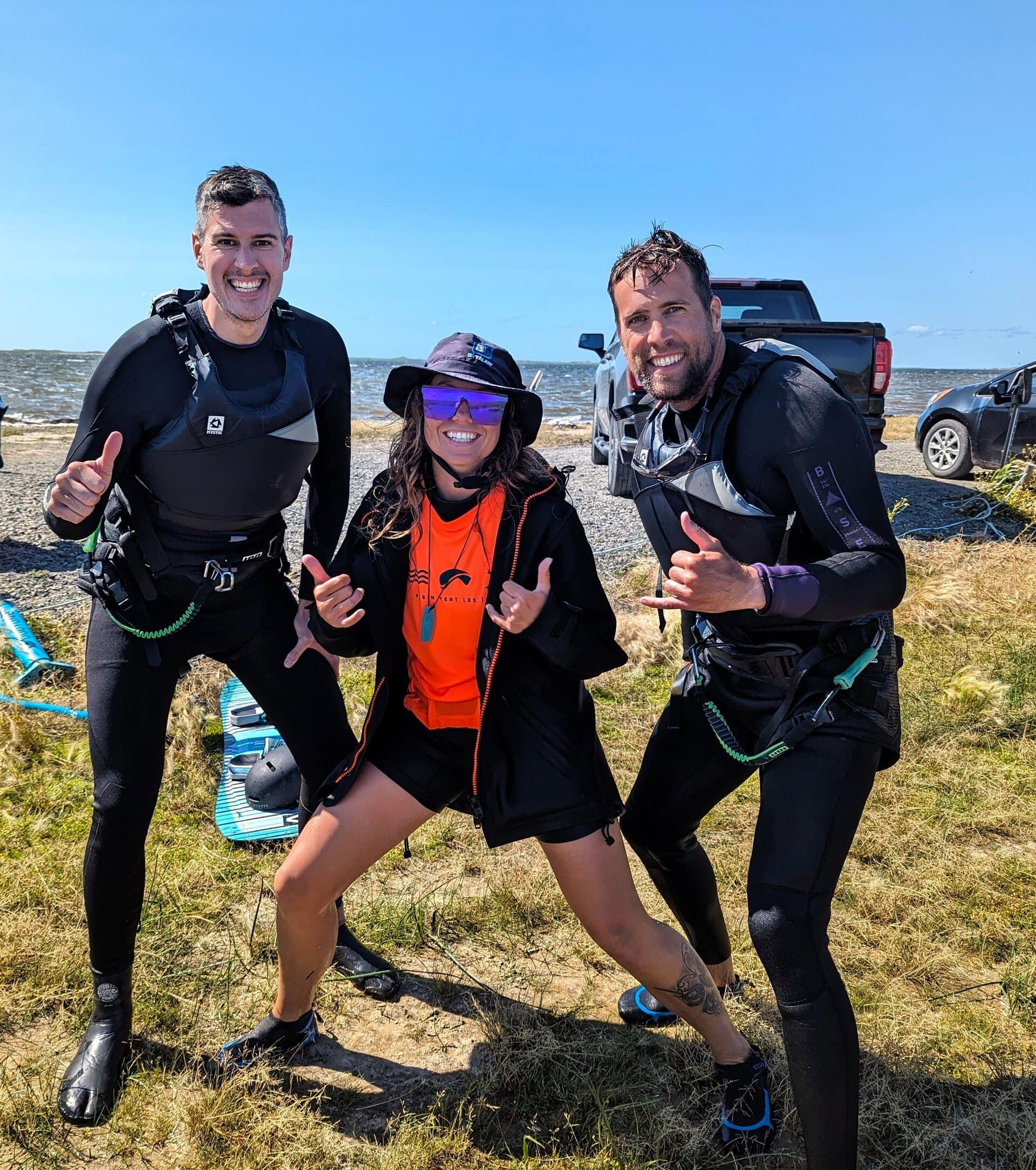 Deux hommes souriants après leur cours de kitesurf en duo, posant avec leur instructrice de Plein Vent les îles.