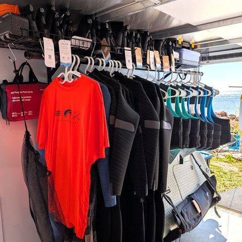 Vue de l'intérieur de la remorque mobile de Plein Vent les îles montrant l'organisation du matériel : combinaisons isothermes (wetsuits) et vestes de flottaison (VFI) prêtes pour les clients.