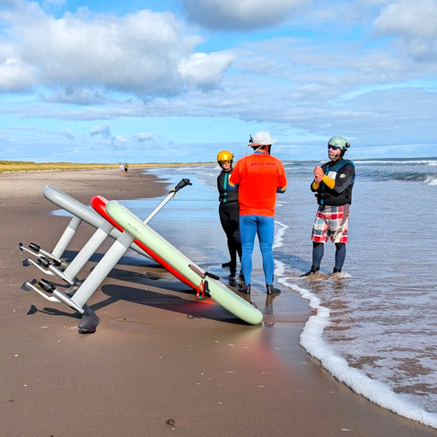 Deux élèves de Plein Vent les îles écoutent la théorie sur la plage. À côté d'eux reposent une planche Fliteboard standard et un FliteScooter, prêts pour la leçon de E-foil aux Îles-de-la-Madeleine.