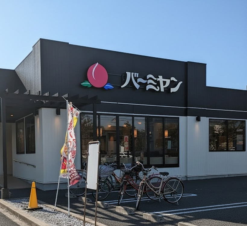 Restaurant entrance with a peach logo and Japanese text. Bikes are parked in front, and a sunny day highlights the black and white exterior.