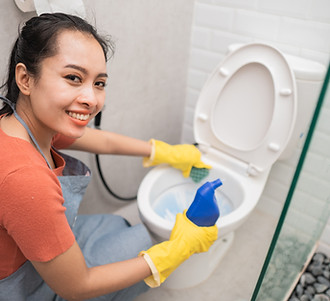 smiling Asian woman wearing gloves holds a bottle of cleaning fluid and a sponge while cle