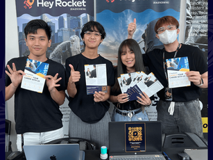 Four Hey Rocket representatives (Ben, Alia, HuiQian, ZhiJie) smiling and holding brochures at a university career fair booth, with branded banners and laptops on display.