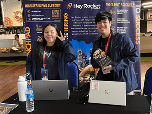 Two Hey Rocket(HonMin Yap and Jessie Kong) representatives standing at their UOW career fair booth, smiling and holding brochures in front of branded banners and laptops.