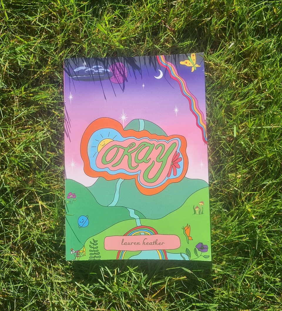 A copy of the book "we are allowed to be okay" laying in the grass. The book cover says the word "okay" in large colourful font, on a background of rolling hills and a prink and purple sunset. There are lots of cartoon images over the top too, such as butterflies and planets and stars in the sky. There is a rainbow across one edge and the authors name "lauren heather"
