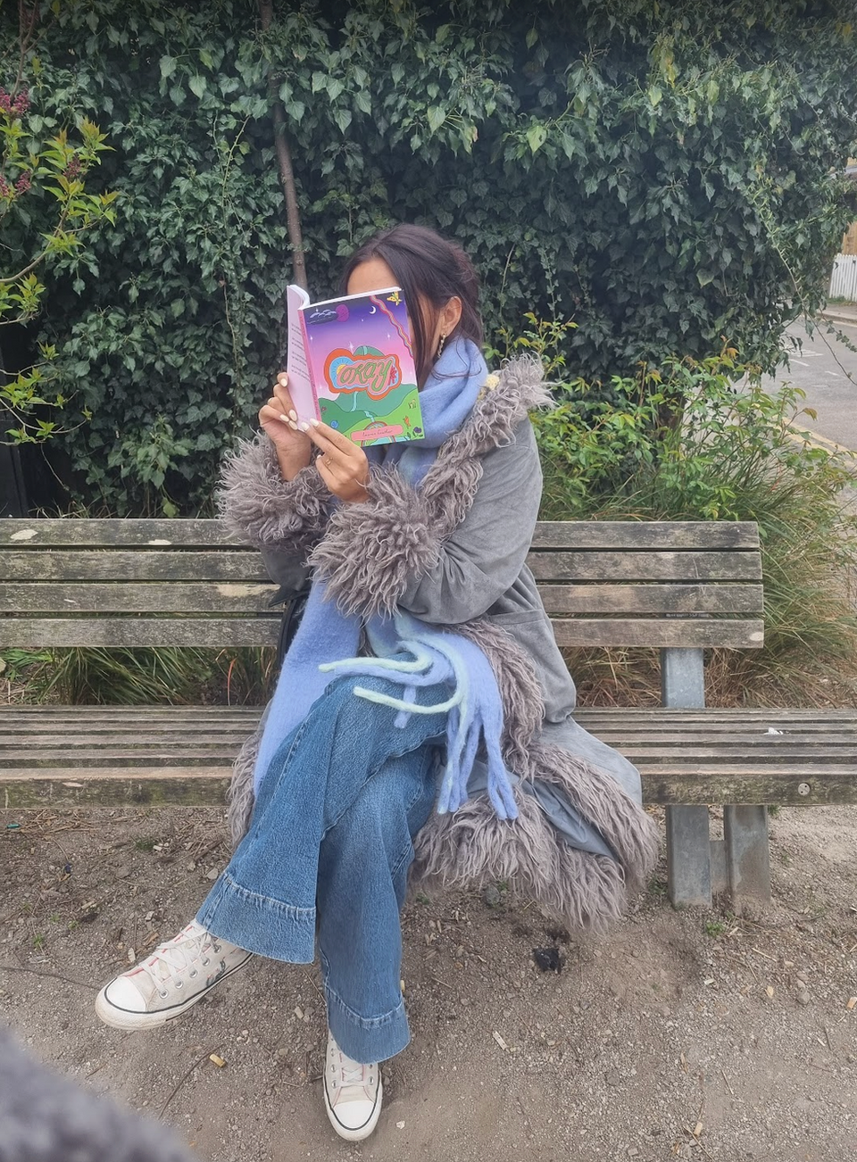 A photograph of the author sat on a park bench, in front of some bushes. The author is a woman wearing an afghan coat, blue jeans and white trainers and she is holding the book open covering her face. You can just see the top of her forehead, and brown hair that is clipped up.
