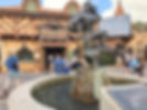 Estátua de personagens da Disney com barris na fonte em frente à Taverna de Gaston, no Magic Kingdom em Walt Dsiney World. Pessoas conversam e passeiam ao redor.