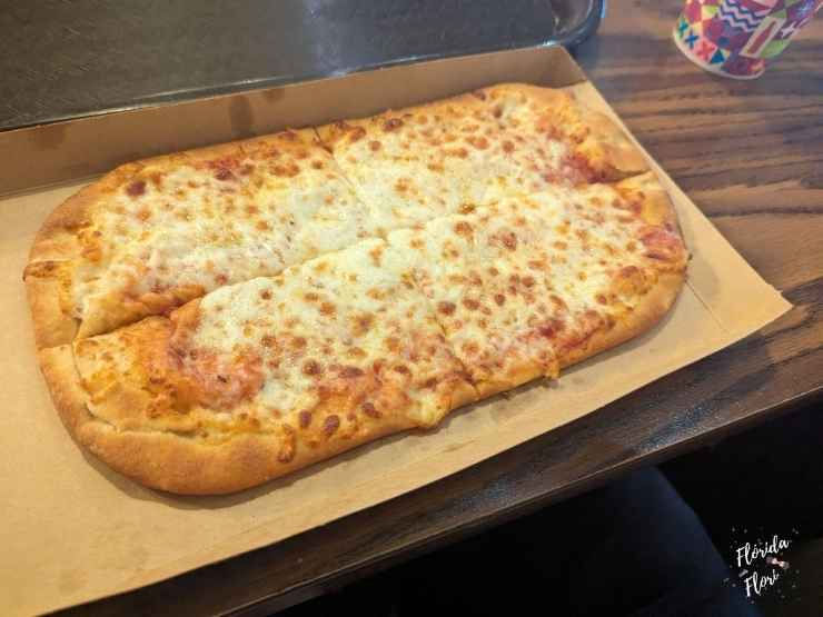 Pizza retangular com queijo derretido sobre bandeja de papel, em mesa de madeira., no restaurante Pinocchio Village Haus, no Magic Kingdom em Walt Disney World. Copo colorido ao fundo. Ambiente descontraído.