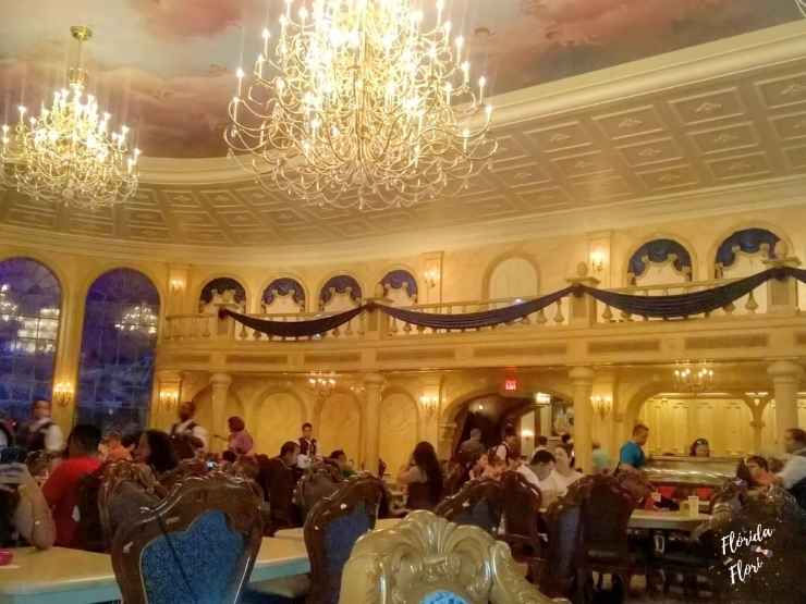 Salão de banquete dourado com candelabros elegantes, pessoas jantando em mesas luxuosas, no castelo da bela e a fera no restaurante Be Our Guest, no Magic Kingdom em Walt Disney World. Ambiente acolhedor e detalhado, luz suave.