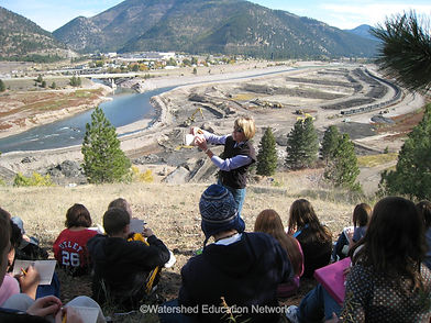 A view overlooking the Milltown Dam site with an instructor standing talking to a group of students sitting on the ground.