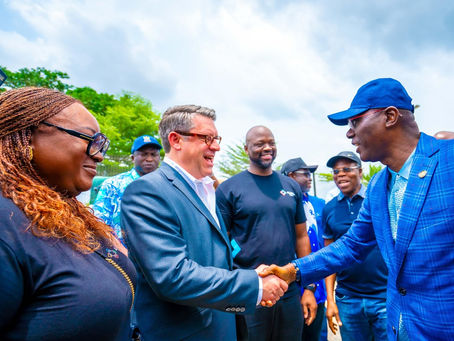 "Lagos State Governor Babajide Sanwo-Olu, wearing a blue checkered suit and cap, shakes hands with a smiling man in a gray suit and glasses during a visit to Alaro City. Several individuals, including a woman in a black top and sunglasses, and officials in blue attire, stand nearby, engaging in conversation under a partly cloudy sky."