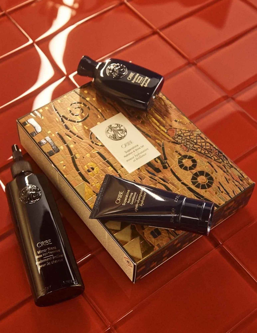 Oribe haircare bottles and tube on a gold patterned gift box atop glossy red tiles, with visible Oribe labels and luxe mood