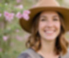 Smiling Mom with fresh healthy skin and a professional haircut in a tan hat amidst blooming pink flowers.