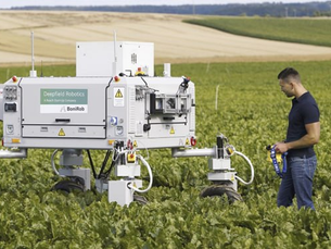 Мужчина стоит в поле рядом с роботом, оба смотрят в сторону горизонта под ясным небом.