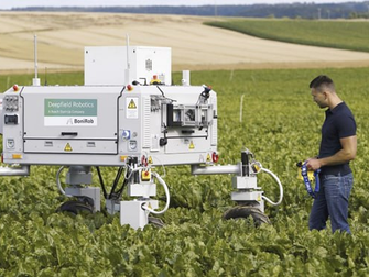 Мужчина стоит в поле рядом с роботом, оба смотрят в сторону горизонта под ясным небом.