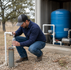 Water testing in progress at a rural Texas residential well system, with a technician filling a sample bottle near visible well equipment.