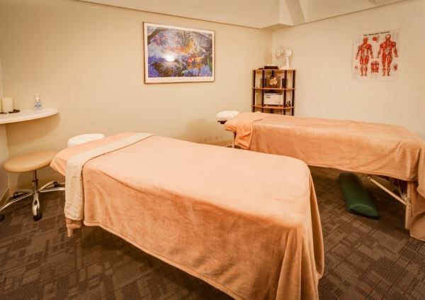 Two massage tables with beige covers in a calm therapy room. Art and anatomy posters on walls; shelves with towels create a serene atmosphere.
