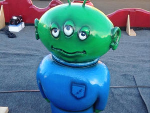 In the image, there is a small or short green alien statue depicted. The alien has three eyes, positioned across its face, two ears protruding from the sides of its head, and two antennae rising from the top of its head. The alien is adorned in a blue jumpsuit, which covers its body. The overall appearance suggests a whimsical and extraterrestrial character, with the statue likely serving as a playful or decorative piece.