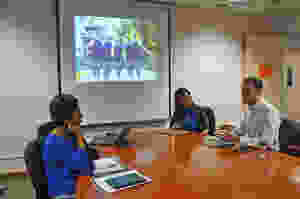 A meeting of three educations around a table. In the background is a PowerPoint slide with an image of the school.