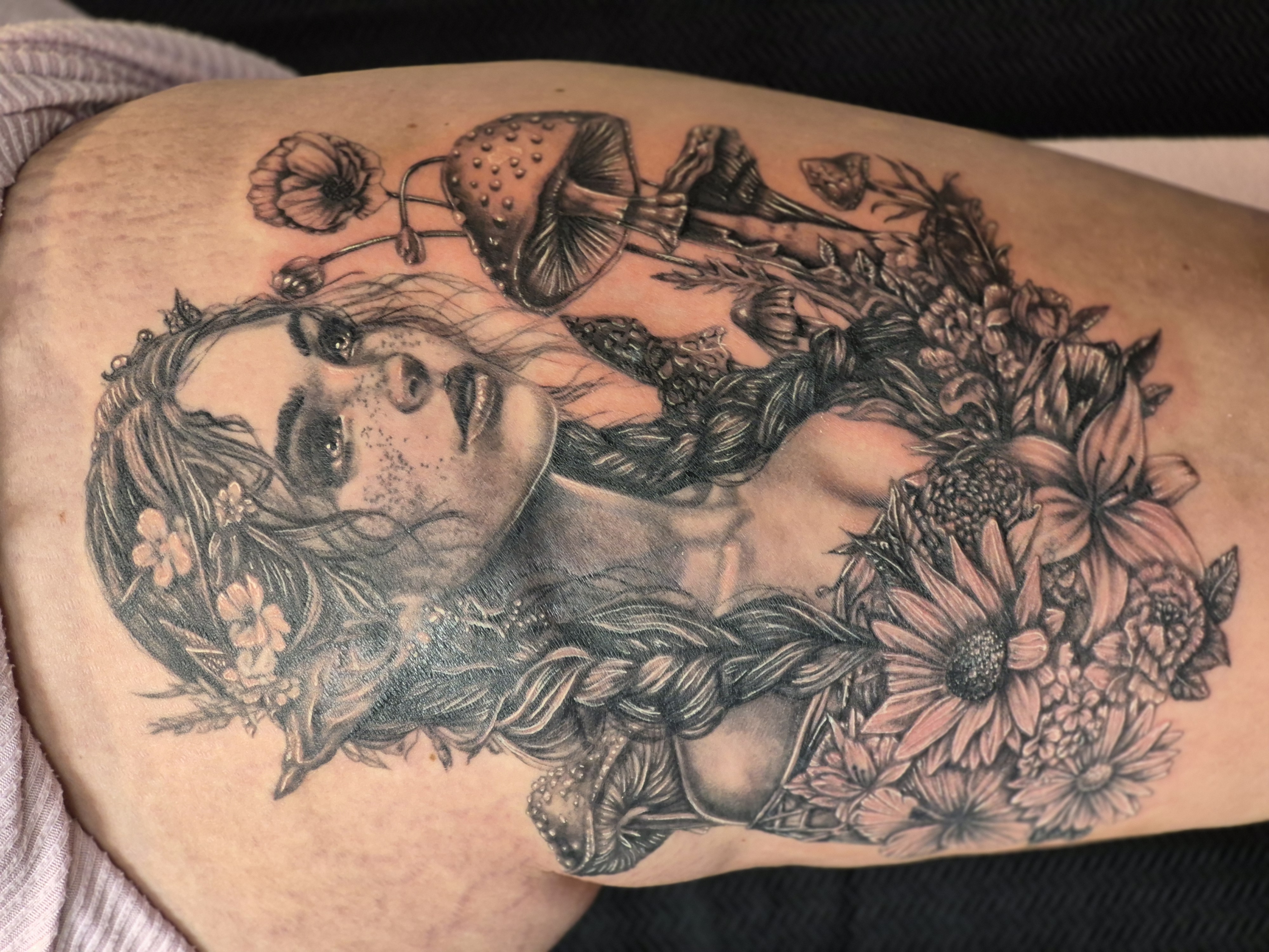 Black-and-grey thigh tattoo of a fantasy woman with pointed ears framed by flowers, mushrooms, foliage, and a small butterfly