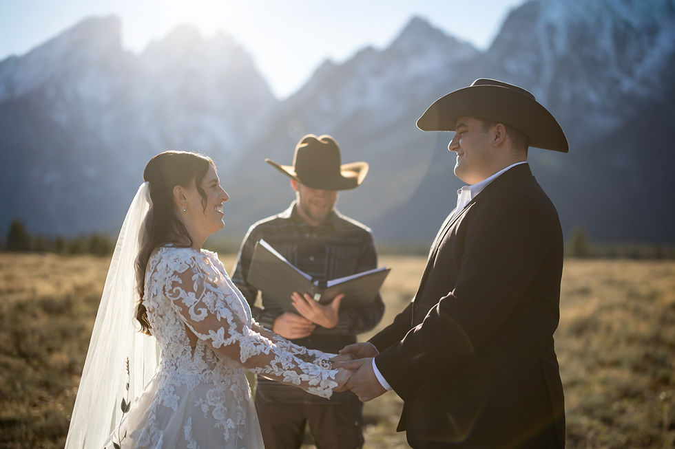 Bride and groom in western attire exchange vows outside with mountains in the background. A smiling officiant holds a book. Sunlit scene.