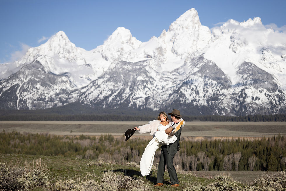 Smiling couple outdoors, groom carrying bride in white dress with the grand teton mountains and blue sky behind them.