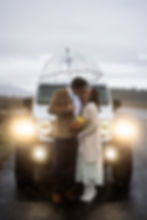Couple kissing under clear umbrella in front of a white SUV with headlights on, holding a bouquet on a rainy road. Moody overcast sky.